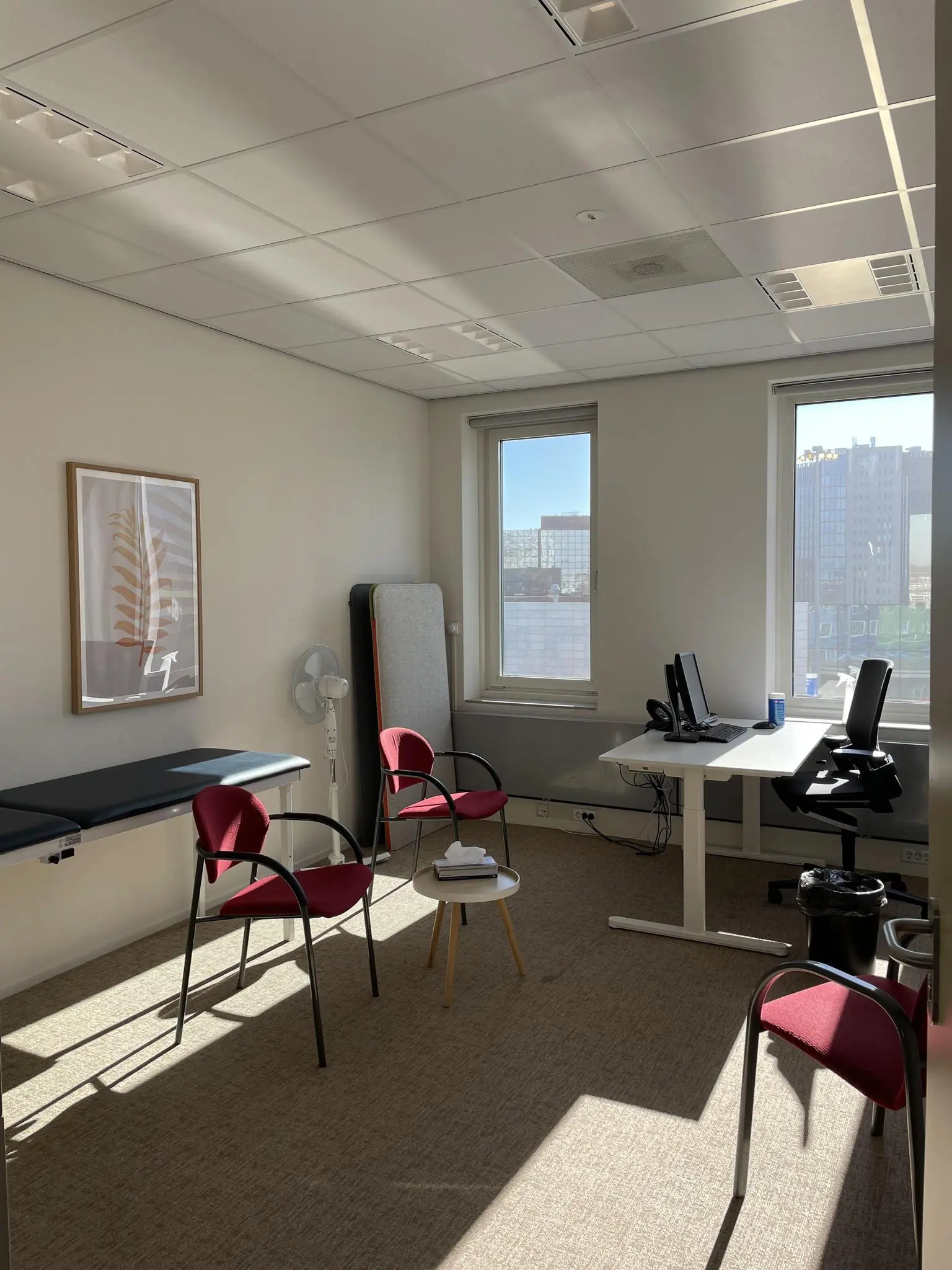 Een modern spreekuur- of behandelkamer op de Johan Cruijff Boulevard met bureau, computer, onderzoeksbank en stoelen in het zonlicht.